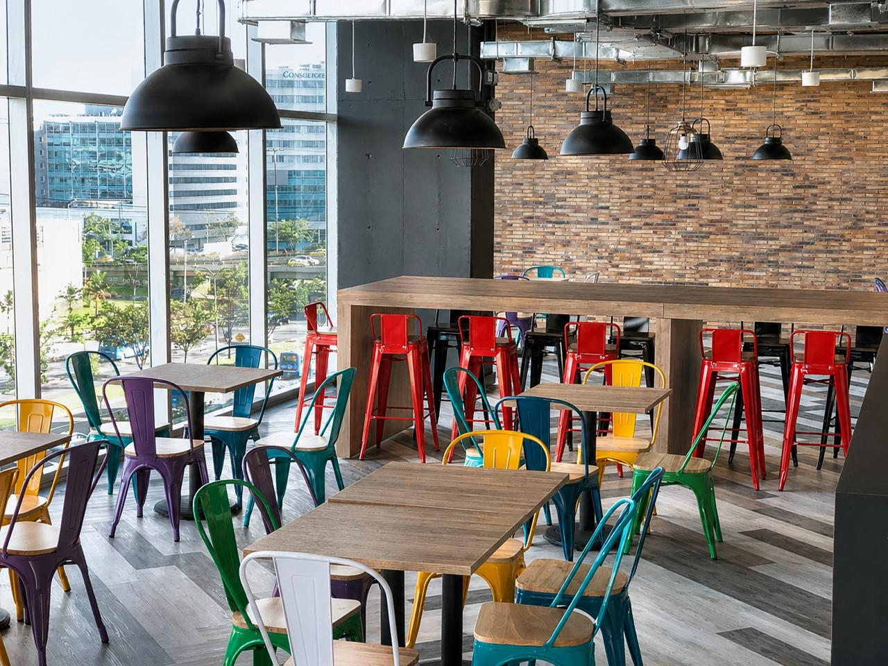 Comedor corporativo con mesón alto de madera, sillas metálicas de colores y muro en ladrillo